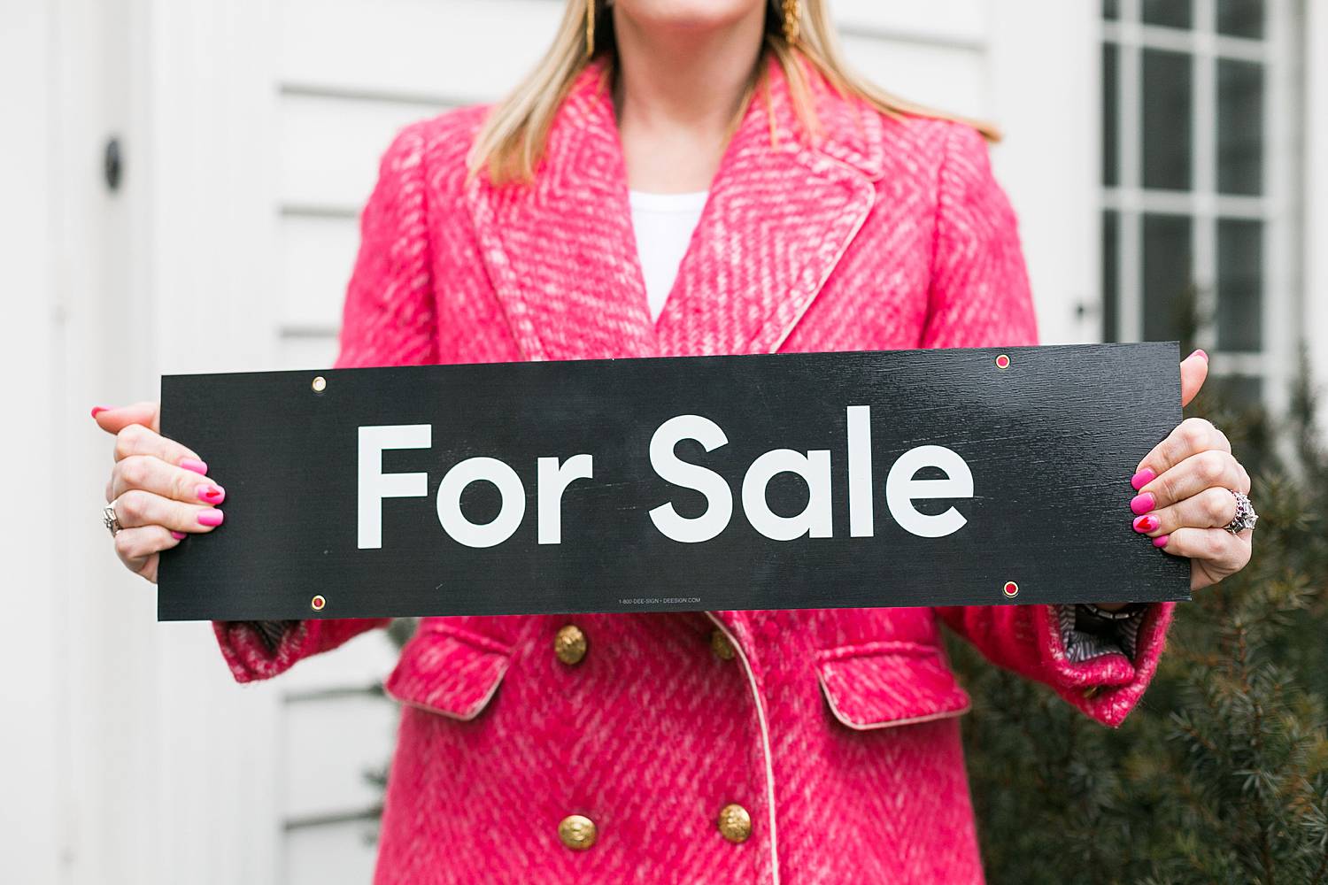 woman real estate agent in pink coat holding sold sign in front of a new home