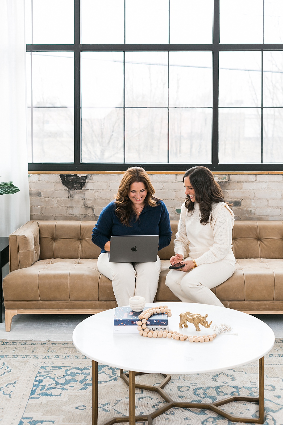 two women working next to each other on a brown leather couch with laptop