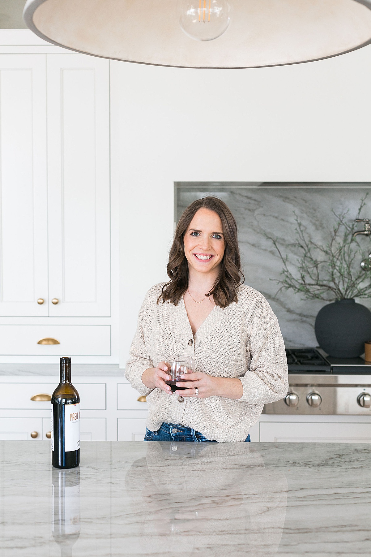woman in tan neutral sweater with a glass of red wine in modern aesthetic kitchen